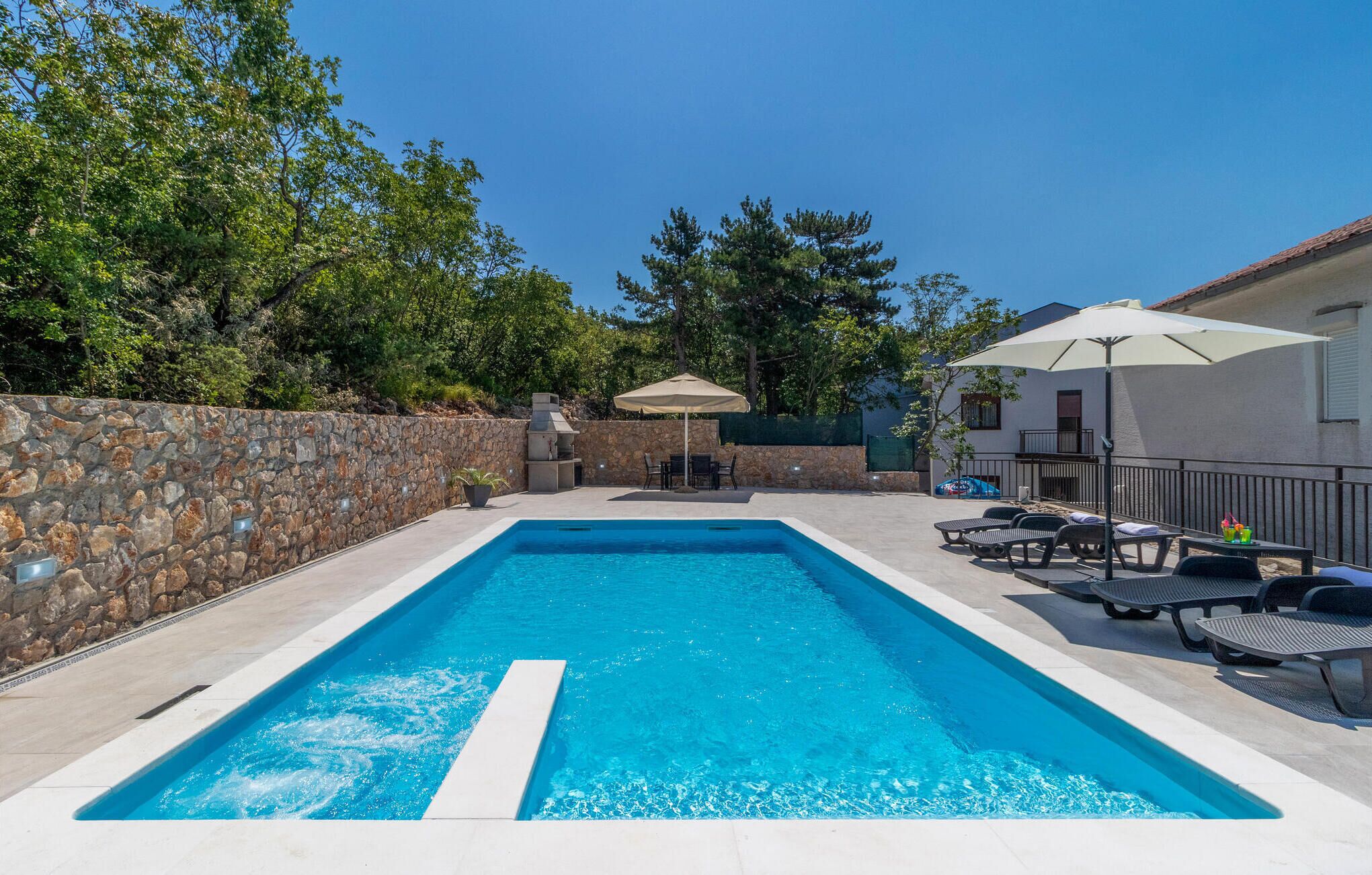 A rectangular swimming pool with a stone wall on one side and a patio area with lounge chairs and umbrellas.