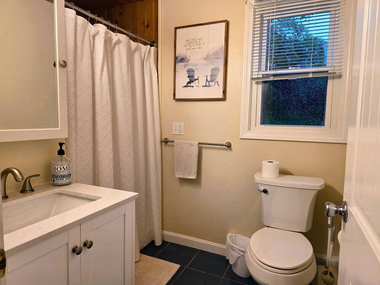 Un baño con lavamanos blanco, inodoro, cortina de ducha y ventana con persianas.