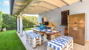 A covered patio with a dining table, two cushioned benches, a refrigerator, and a kitchen area with a sink and a fish-shaped decoration. - Beach front home in Casuzze-Finaiti with sauna (Casuzze-Finaiti)