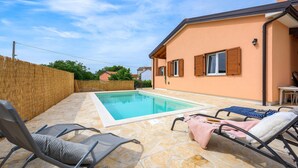 Un espace piscine aménagé avec des chaises longues et une table, entouré d’une terrasse en pierre et d’une clôture en bois.