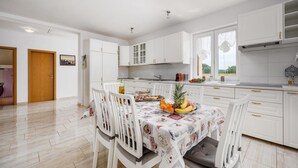 Une cuisine moderne avec des meubles blancs, une table à manger avec des chaises et une vue sur l’extérieur grâce à de grandes fenêtres.