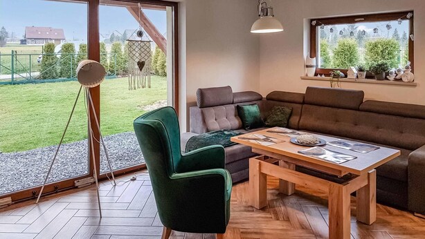 Ein modernes Wohnzimmer mit einer Couch, einem grünen Sessel, einem Holztisch und einem großen Fenster mit Blick auf eine grüne Wiese und ein Haus.