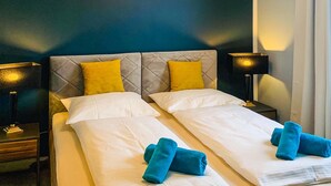 Uma cama de casal com lençóis brancos e duas toalhas azuis, um encosto estofado e duas luminárias de cabeceira.