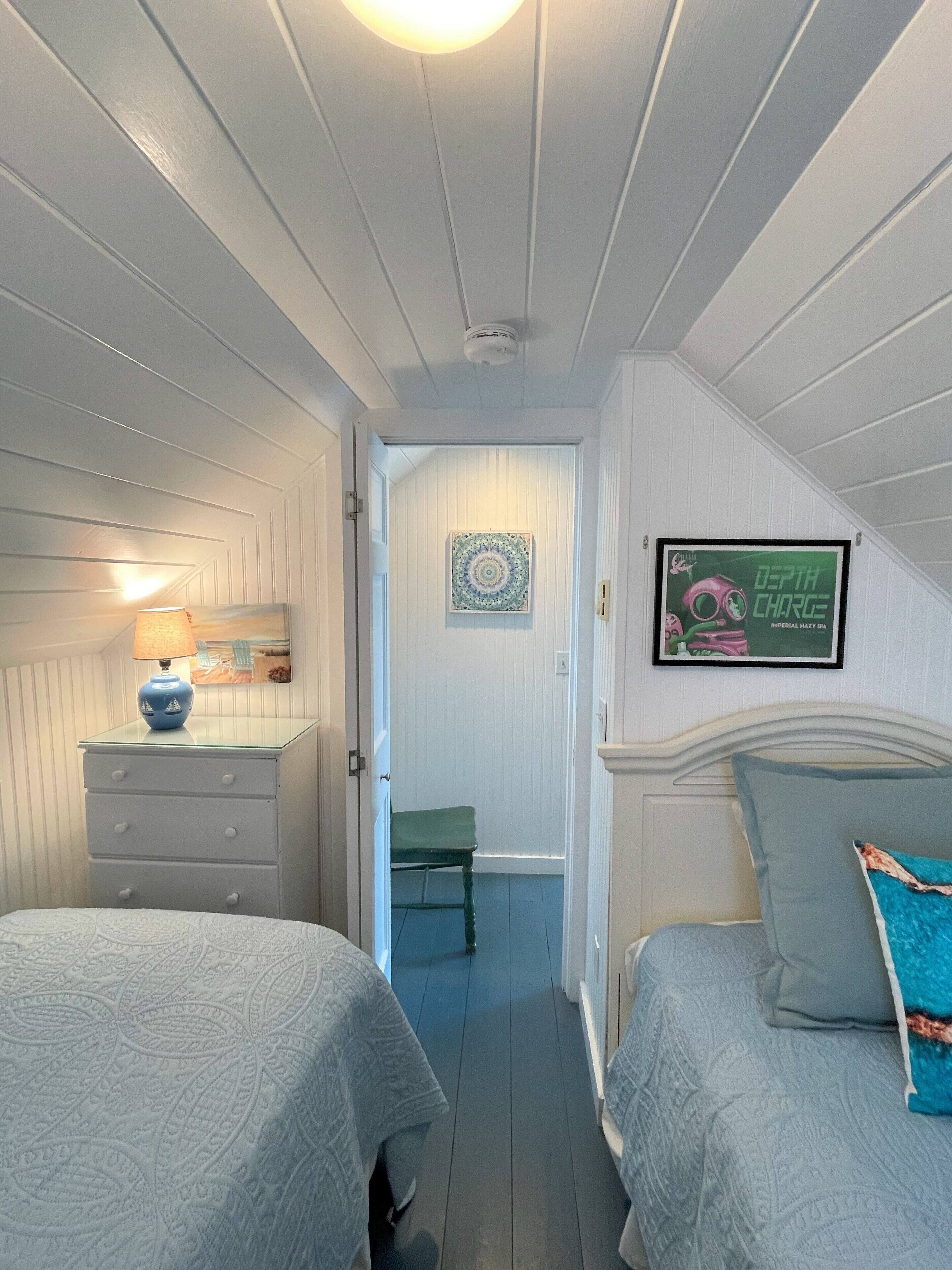 A bedroom with two beds, a dresser, a lamp, a framed picture, and a door leading to another room.