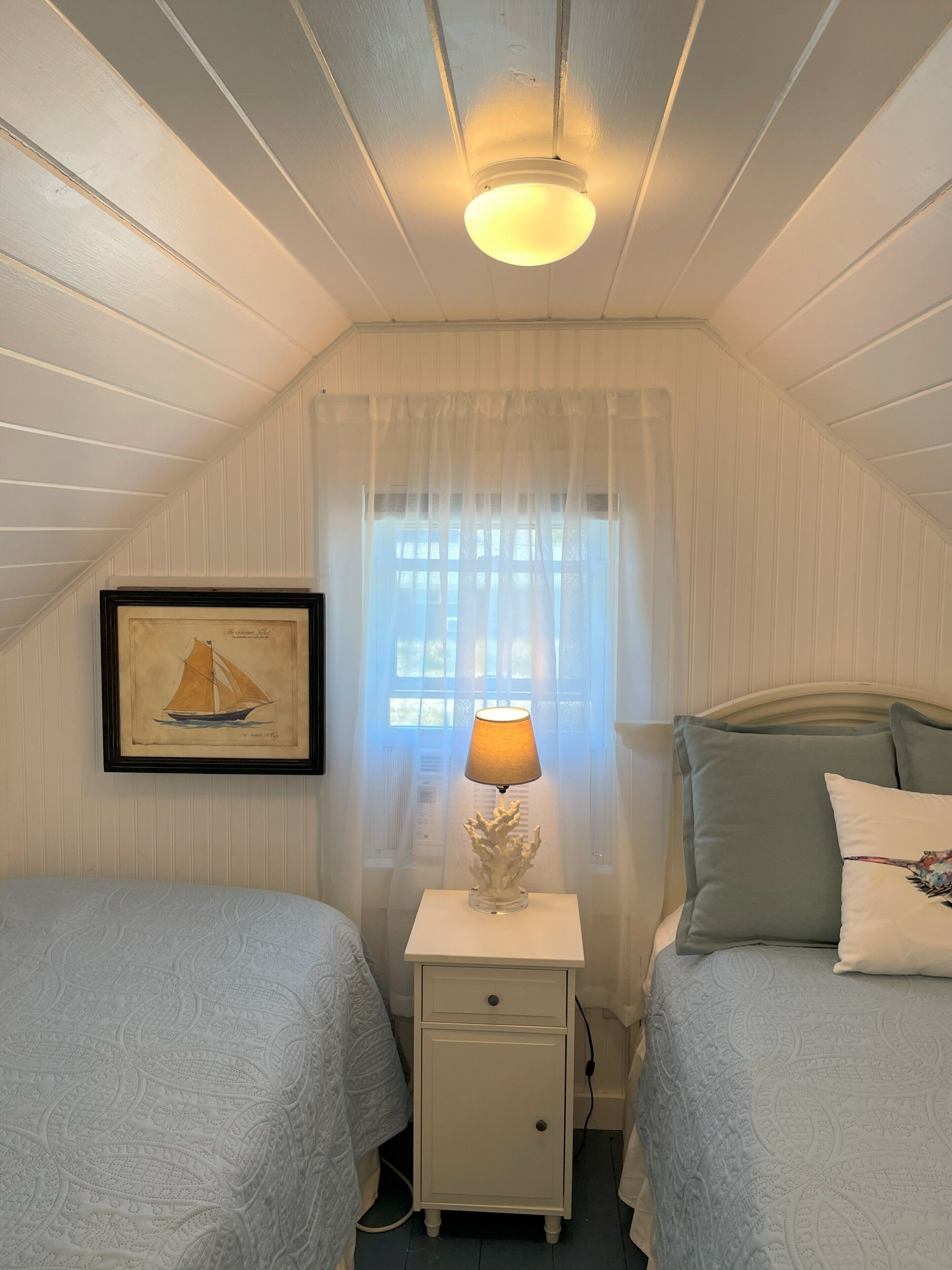 A bedroom with two beds, a nightstand with a lamp, a framed picture on the wall, and a window with sheer curtains.