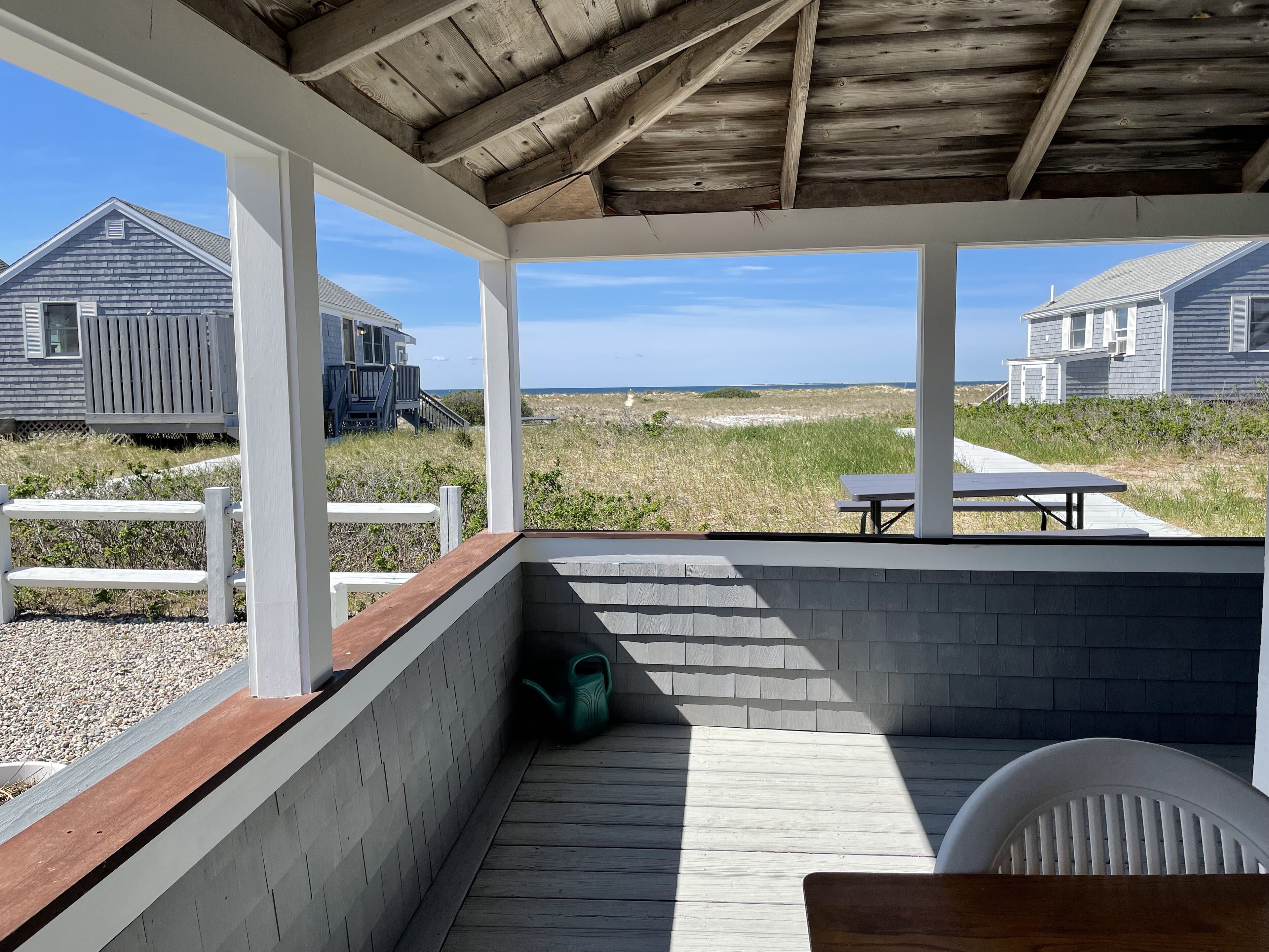 A covered outdoor area with a wooden deck, white railings, and a view of the beach and houses.