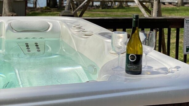 A hot tub with a bottle of wine and a glass, surrounded by a wooden deck and trees, overlooking a body of water. - Lakefront, Wooded, & Secluded Home - Family Dogs Welcome. (Oakland)