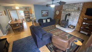A cozy living room with a blue sofa, a brown leather ottoman, and a fireplace. There is a dining area with a wooden table and chairs, and a kitchen in the background. - Lakefront, Wooded, & Secluded Home - Family Dogs Welcome. (Oakland)