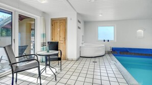 A poolside area with a glass door leading to a balcony, a round table with chairs, a wooden door, a bathtub, and a window overlooking the pool.