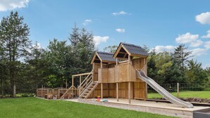 A wooden play structure with a slide and stairs, surrounded by grass and trees.