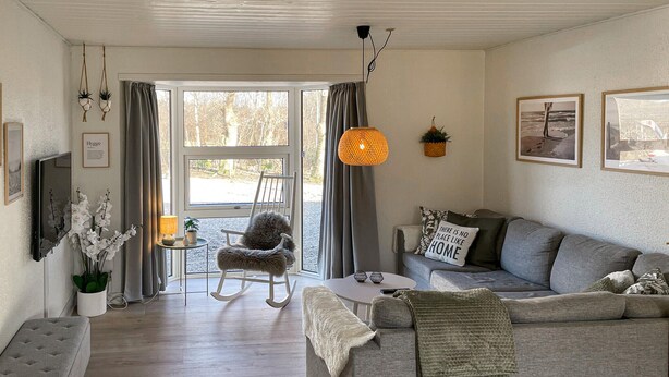 A cozy living room with a grey sofa, a white armchair, and a small table with a lamp. There are two hanging plants, a framed picture on the wall, and a window with curtains.