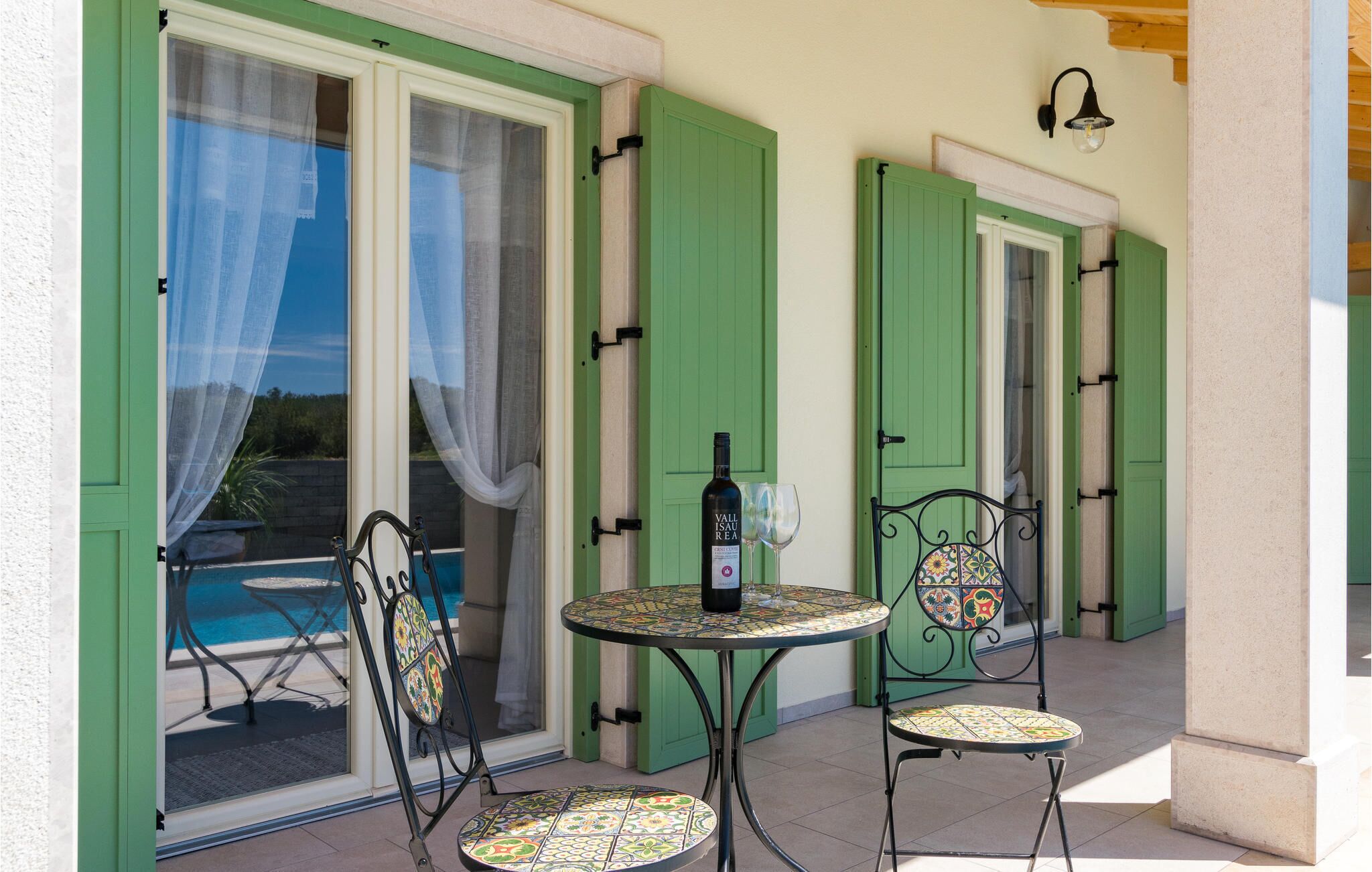 A patio with a pool, green shutters, a table with a bottle of wine, and two chairs.