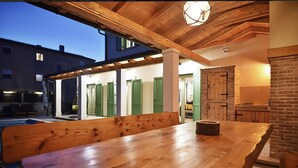 An outdoor wooden table with benches, a brick wall, and a building with green doors in the background. - Gorgeous home in Peroj (Peroj)