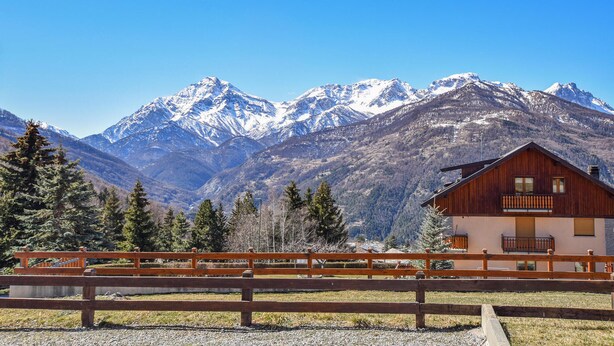 A wooden cabin with a balcony, surrounded by a wooden fence, set against a backdrop of snow-capped mountains and evergreen trees.