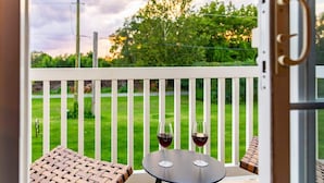 Una terraza con una mesa y dos sillas, una copa de vino tinto sobre la mesa, y vistas a un césped verde y árboles.