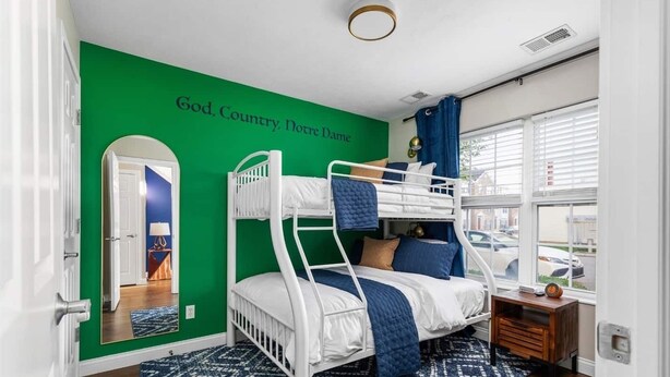 Ein Zimmer mit Etagenbetten, einer grünen Wand mit einem Schriftzug, einem Fenster mit Jalousien und einer Tür mit Spiegel.