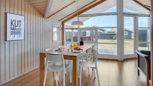 Un comedor con mesa de madera y sillas blancas, un ventanal con vista a la playa y un cartel en la pared.