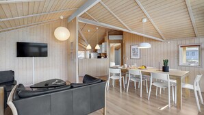 Un interior moderno con techo de madera, un comedor con sillas blancas, un sofá de cuero negro y una cocina visible a través de un vano abierto.