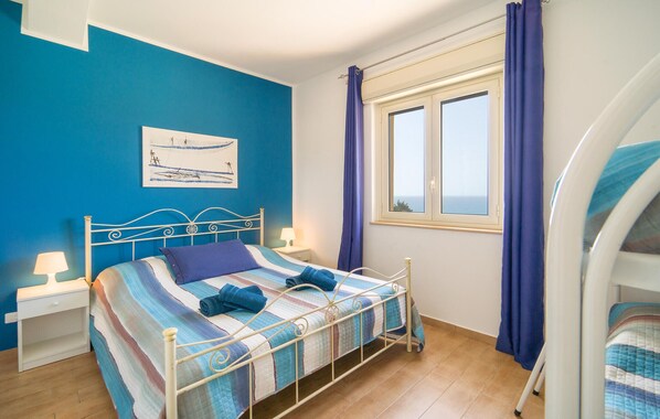 Een slaapkamer met een bed, nachtkastje, lamp, raam met gordijnen en uitzicht op zee.