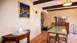 Un comedor con mesa de vidrio, sillas de madera, una mesita con jarrón y platón, un cuadro de un paisaje urbano y una araña.