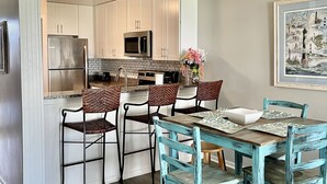 A modern kitchen with white cabinets, stainless steel appliances, and a dining area with mismatched chairs and a rustic table. - Tommy's Bahama! in A Place At The Beach Resort on the OCEAN & the BEST pools! (Atlantic Beach)