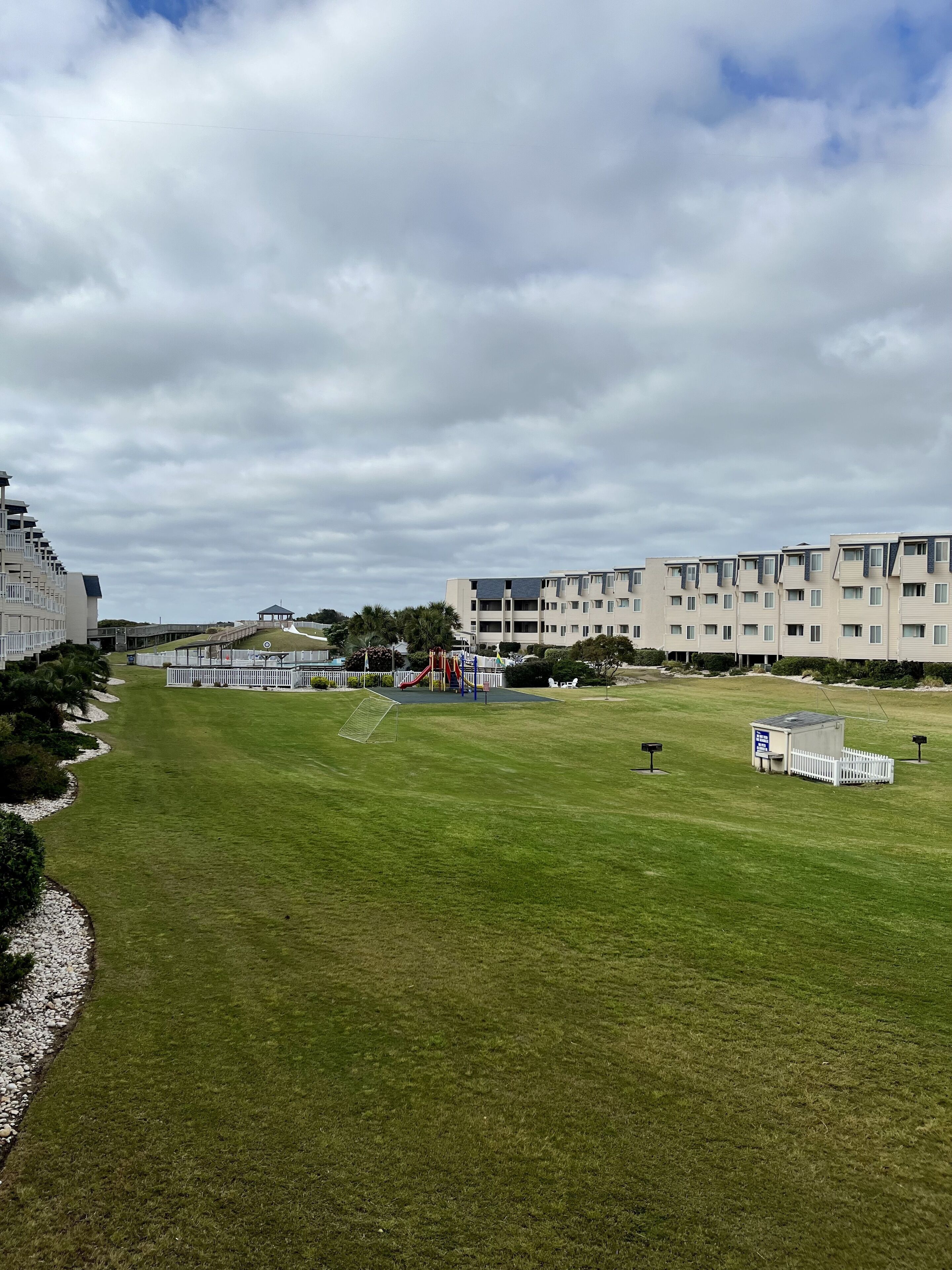 Un campo cubierto de césped con juegos infantiles, una cerca blanca y modernos edificios de apartamentos al fondo.