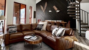 Ein geräumiges Wohnzimmer mit einer großzügigen L-förmigen Eckcouch, einem runden Couchtisch und einem schwarz-weißen Streifen Teppich. Eine dunkle Holz-Akzentwand ziert die Räumlichkeit mit dekorativer Wandkunst, und eine Treppe mit einer einzigartigen Steinreling führt nach oben.