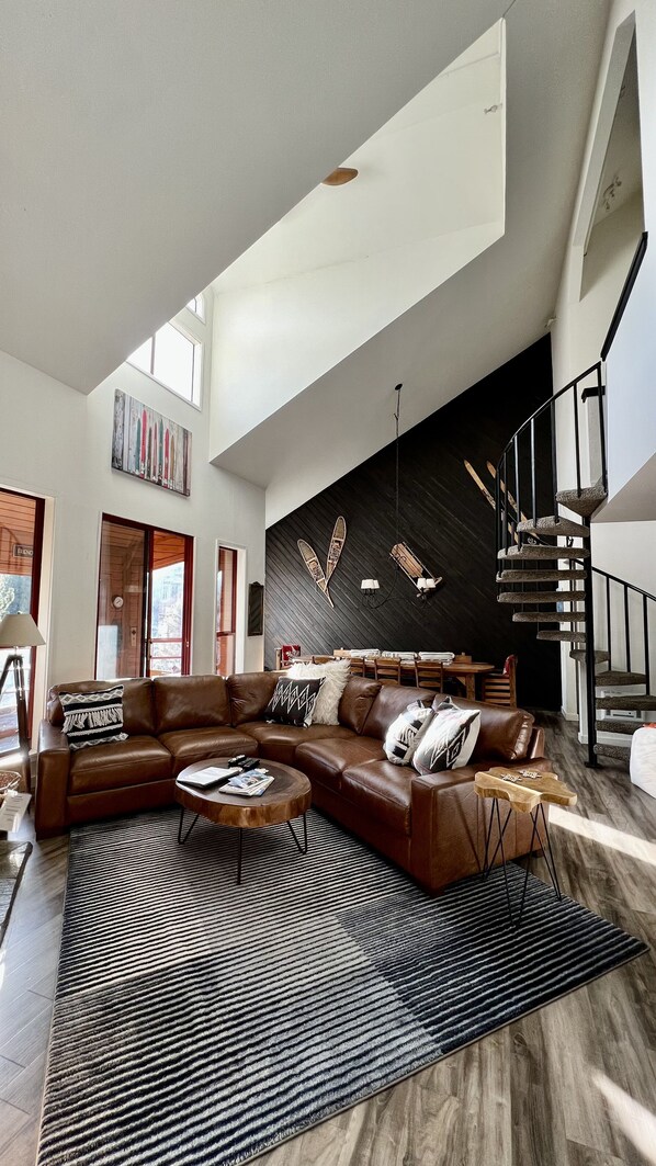 A spacious living room with a large L-shaped sectional sofa, a round coffee table, and a black and white striped rug. There is a dark wooden accent wall with decorative wall art and a staircase with a unique stone railing.