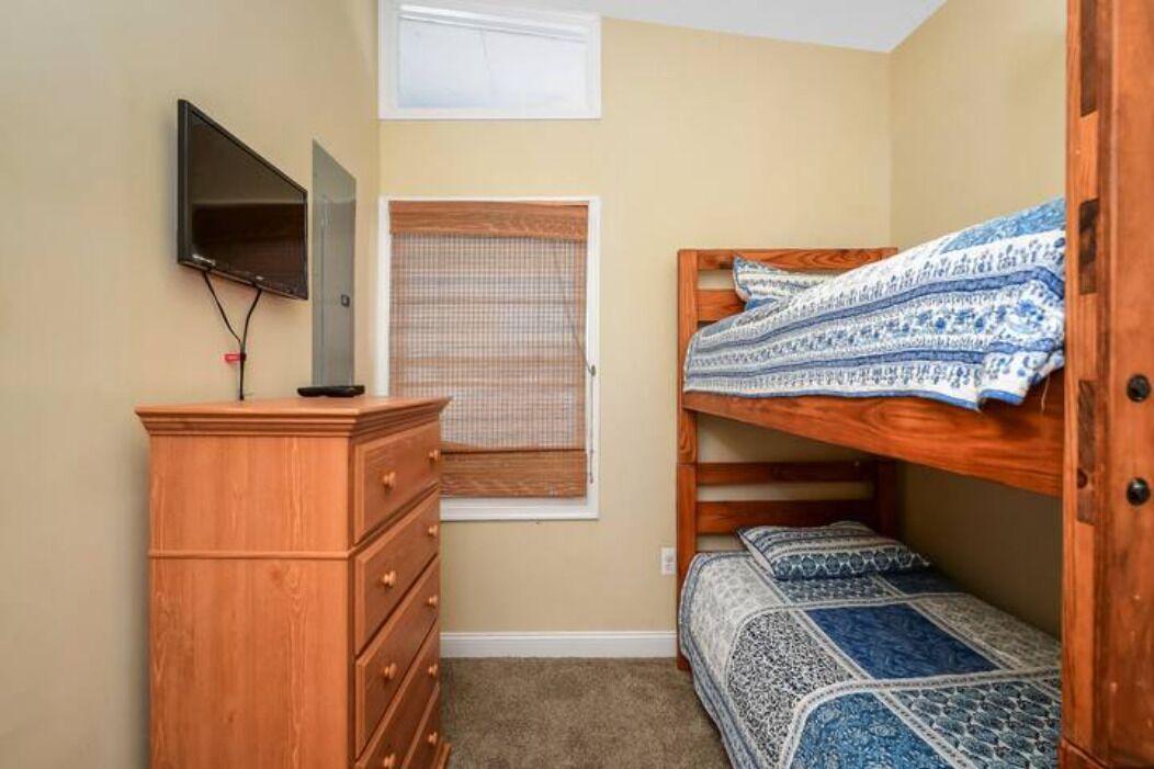 A bedroom with a bunk bed, a wooden dresser, a television mounted on the wall, and a window with a blind.