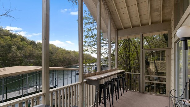 A covered outdoor area with a view of a lake and trees. There are several black bar stools and a white table. - Lake front property with huge dock connected to Margaritaville (Osage Beach)