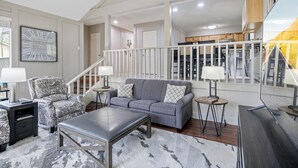 A spacious living room with a grey sofa, patterned armchair, and a coffee table. A staircase with a railing leads to an upper level. - Lake front property with huge dock connected to Margaritaville (Osage Beach)