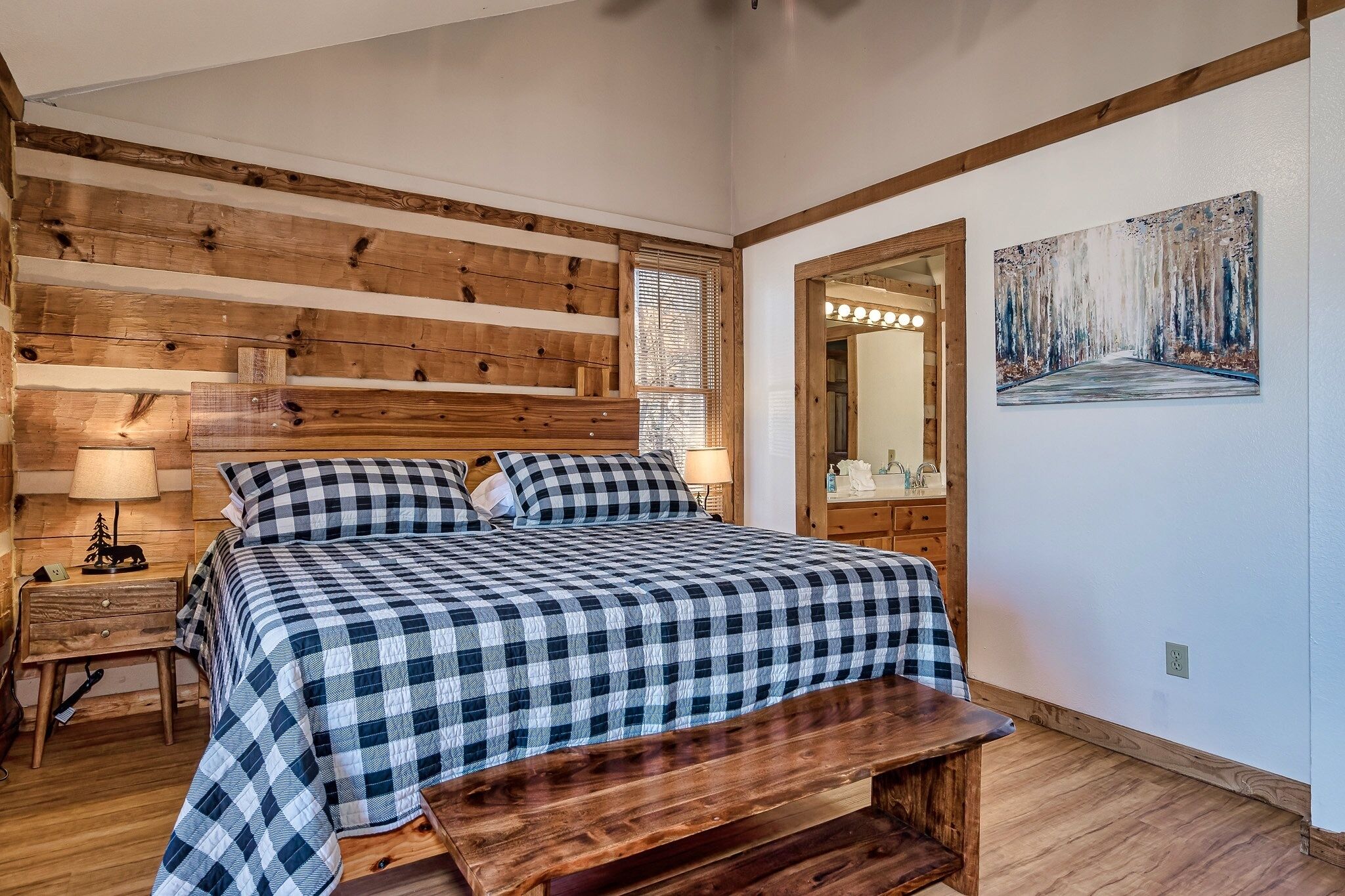 A bedroom with a wooden headboard, a checkered bedspread, a bedside table with a lamp, and a bench.