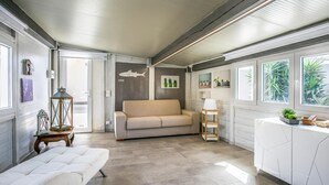 A compact, well-lit room with a sofa, a small table, and a cabinet. There are windows with plants and a wall with a shark decal. - Gorgeous home in Santa Croce Camerina (Santa Croce Camerina)