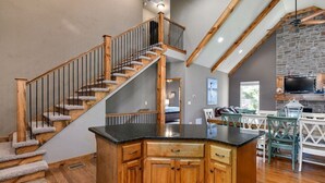 A spacious kitchen with a central island, wooden staircase, and a stone fireplace.