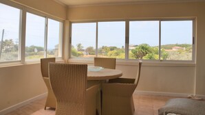 Una habitación con una mesa redonda y cuatro sillas de mimbre, ventanas amplias con vistas a zonas verdes y edificios, y suelo de baldosas.