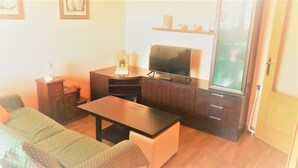 Un espace de vie compact comprenant un canapé, une table basse, un téléviseur posé sur un meuble et une petite étagère agrémentée d’objets décoratifs.