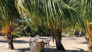 A beach setting with a wooden table and two chairs under a palm tree, surrounded by sand and other vegetation. - La Vida Belize - Casa is a Beautiful Beach Front Home with Pool (Placencia)
