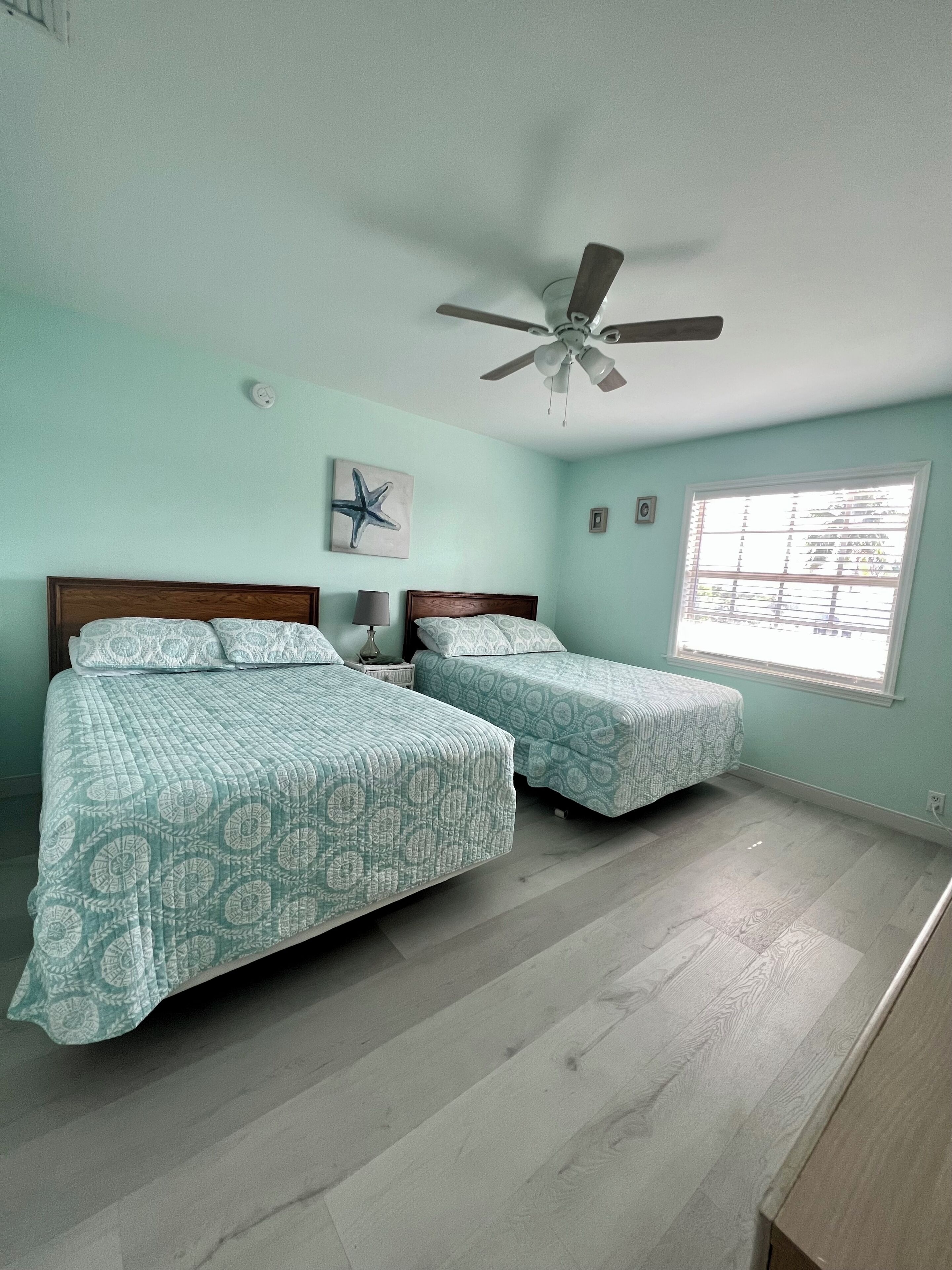 A bedroom with two beds, a ceiling fan, a window with blinds, and a starfish artwork on the wall.