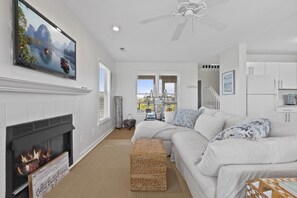 A modern living room with a fireplace, white sofas, a large flat-screen TV, and a ceiling fan.