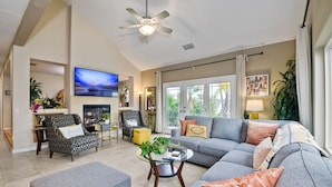 A spacious living room with a grey sofa, ottoman, and a glass coffee table. A television is mounted on the wall, and there is a ceiling fan.