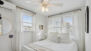 Ein Schlafzimmer mit Bett, Deckenventilator, Fenster mit Vorhängen, Nachttisch und Wanduhr.
