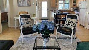 Una sala de estar con dos sillones, una mesa de centro y un jarrón de flores. Se observa una cocina al fondo, con un refrigerador y armarios.