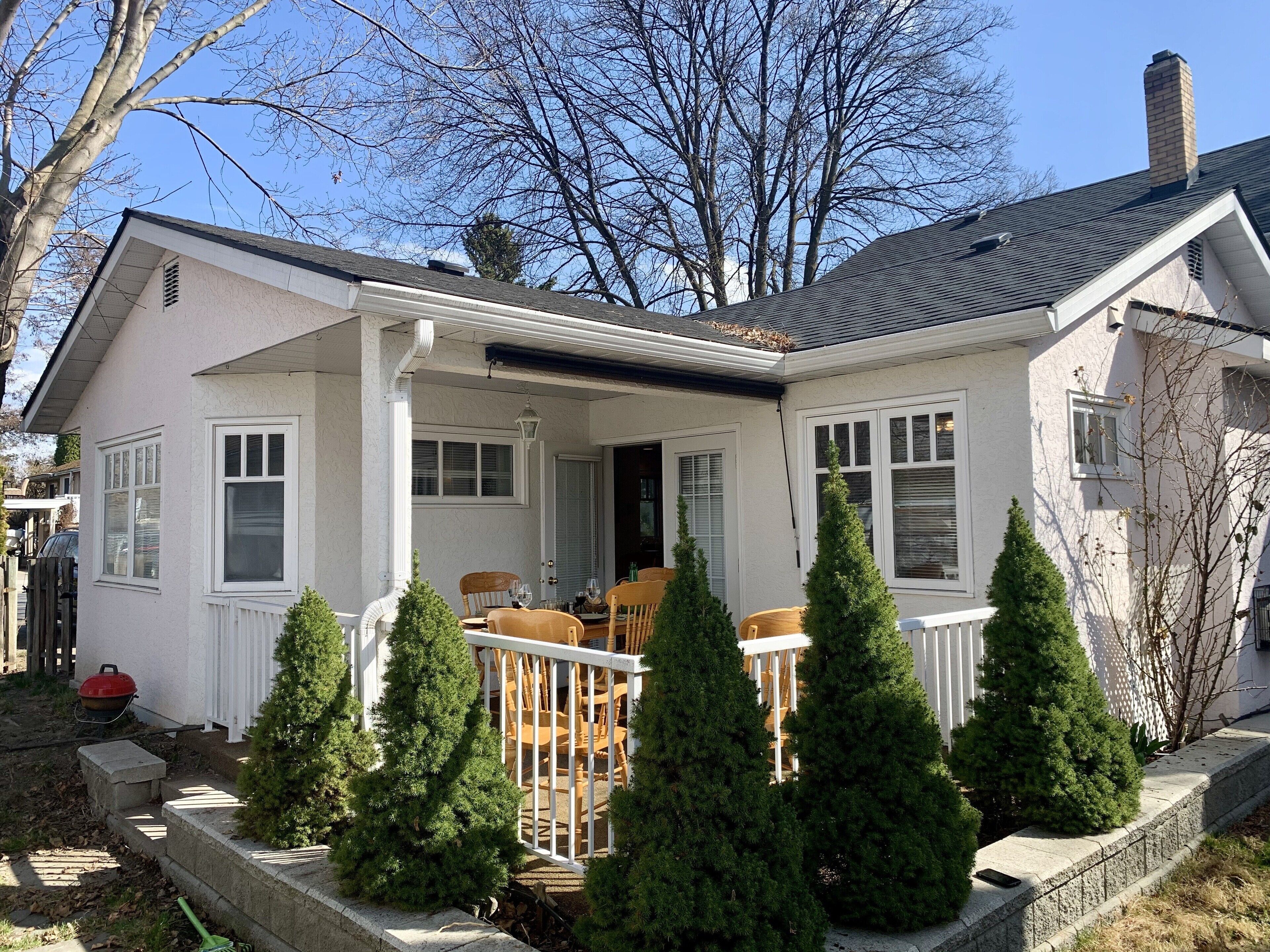 Ein zweistöckiges Haus mit einer Veranda, einem Schornstein und einem kleinen Garten mit immergrünen Bäumen.