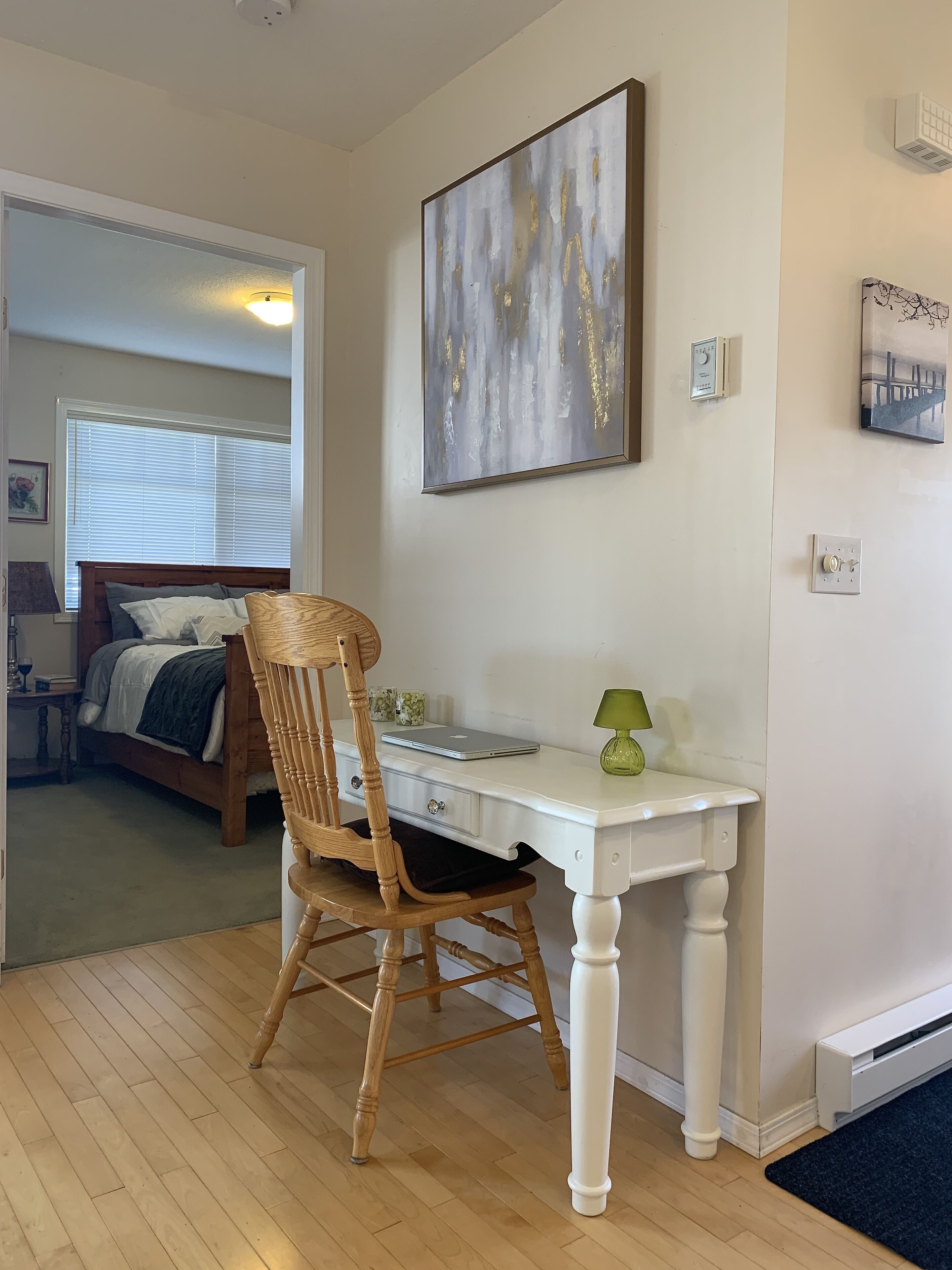 Una habitación con una silla de madera, un escritorio blanco, una computadora portátil y un cuadro en la pared.