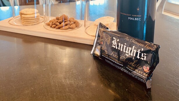 Una botella de vino Malbec Moonlight Harvest, un paquete de chocolate oscuro Knight's, y una bandeja con galletas y almendras sobre una mesa de comedor en una sala de estar.