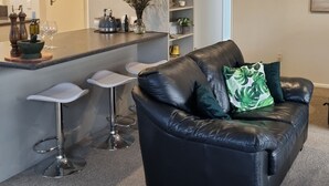 Uma sala de estar moderna com um sofá de couro preto, um pequeno espaço de bar e uma cozinha ao fundo.