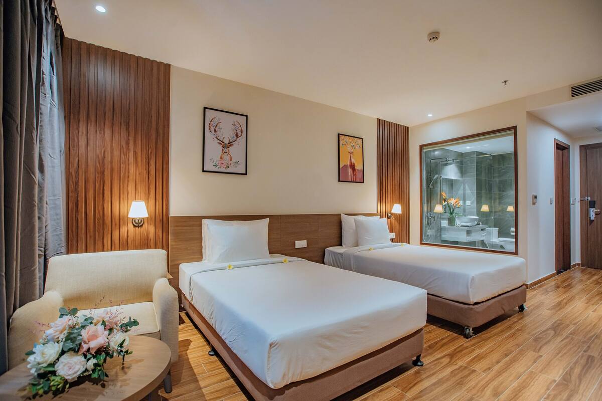 Een hotelkamer met twee bedden, een houten hoofdbord, een klein tafeltje met een bloemstuk en een groot raam met uitzicht op een badkamer en een deur.
