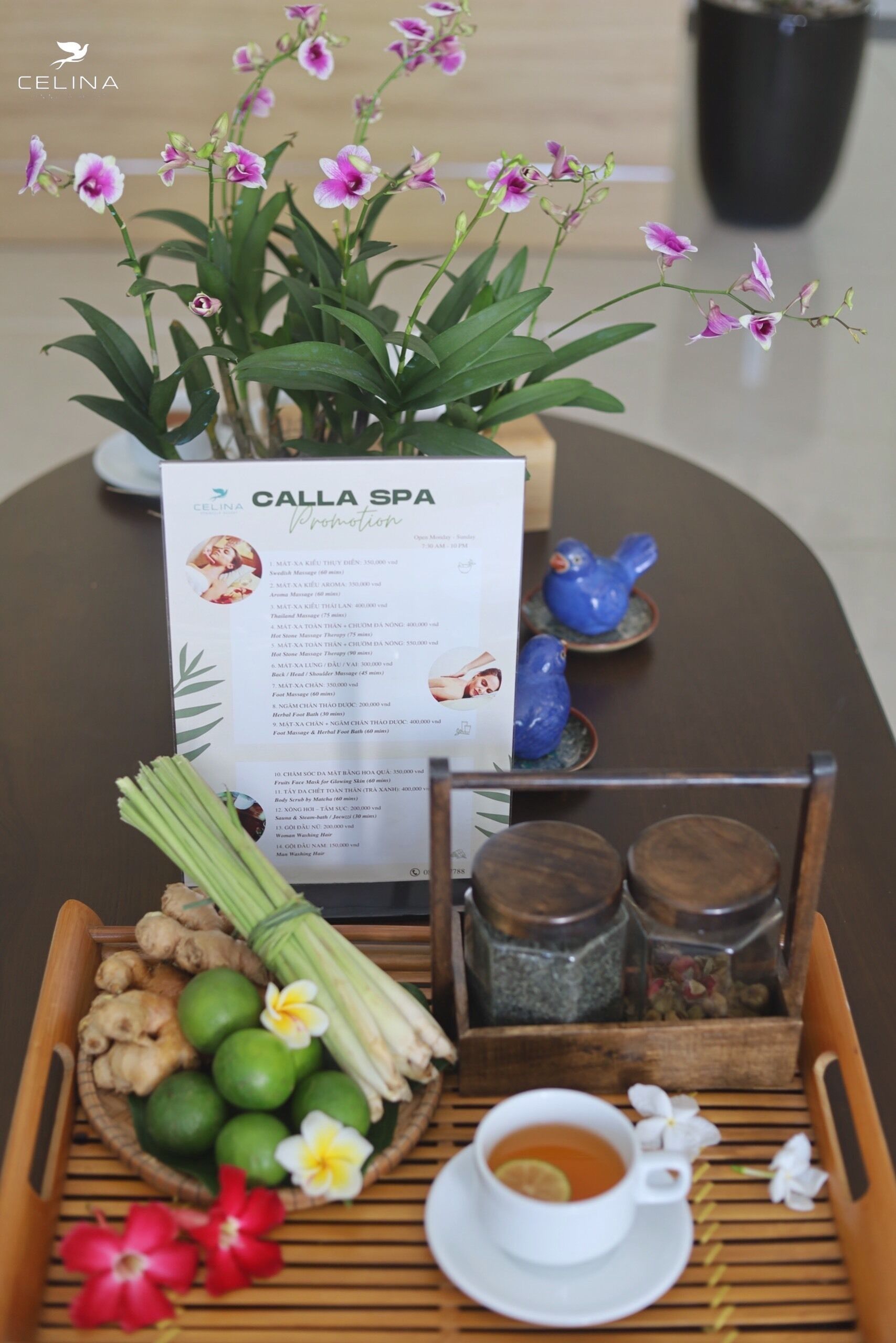 A table with a menu, a tray of ginger, lime, and flowers, a cup of tea, and a potted orchid plant.