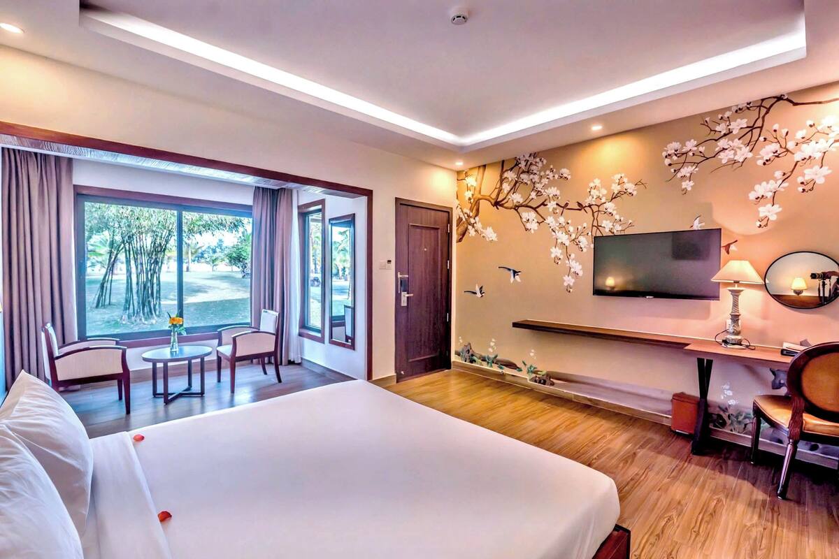 Een hotelkamer met een groot bed, een televisie, een bureau met een lamp, een stoel en uitzicht op bomen door het raam.