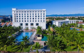 A large hotel with a central swimming pool surrounded by palm trees and a view of the cityscape. - Celina Peninsula Resort (Dong Hoi)